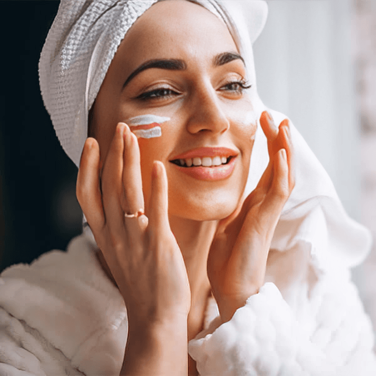 A person with a towel on their head applying cream to their face.