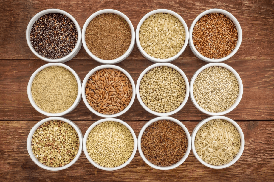 Twelve bowls of various grains on a wooden table, top view.