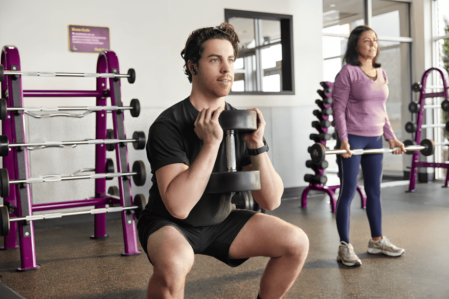 A man squats with a dumbbell; a woman stands nearby.