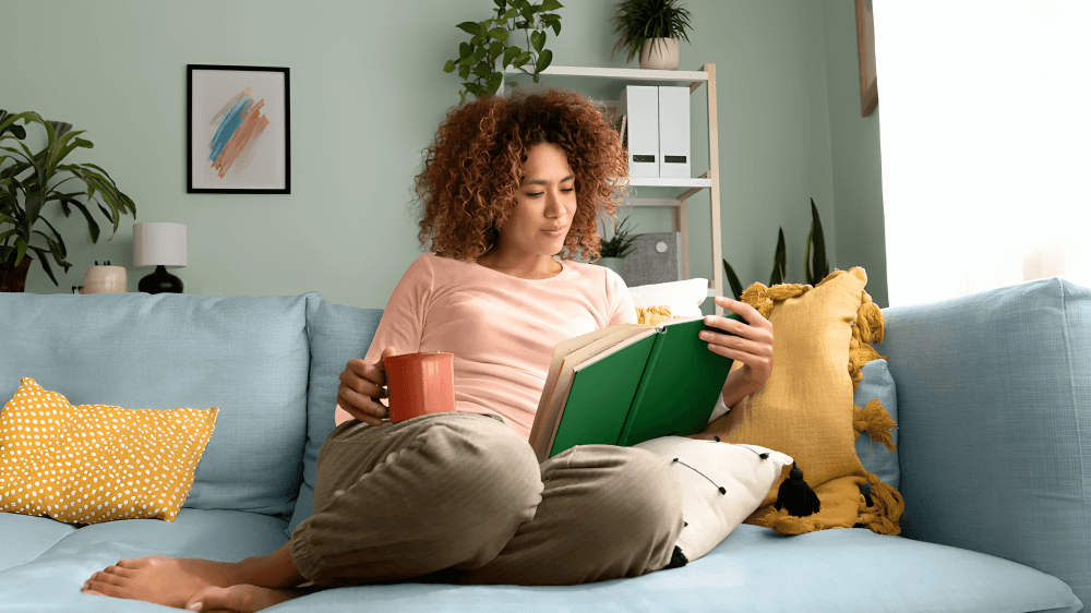 A curly-haired woman reads a book on a sofa.