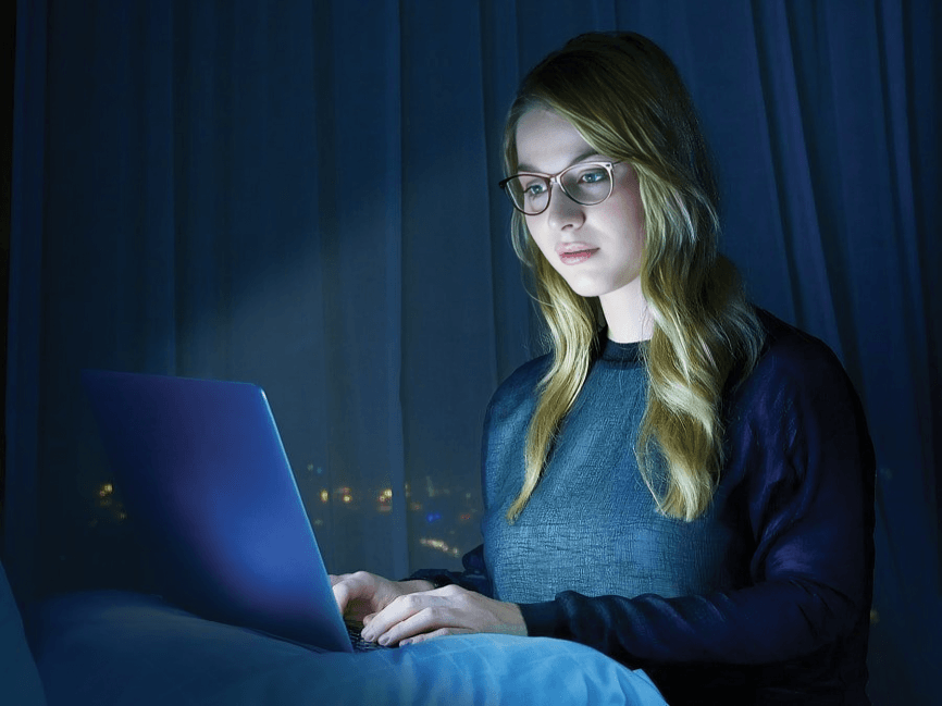Woman with glasses using laptop in dim bedroom.