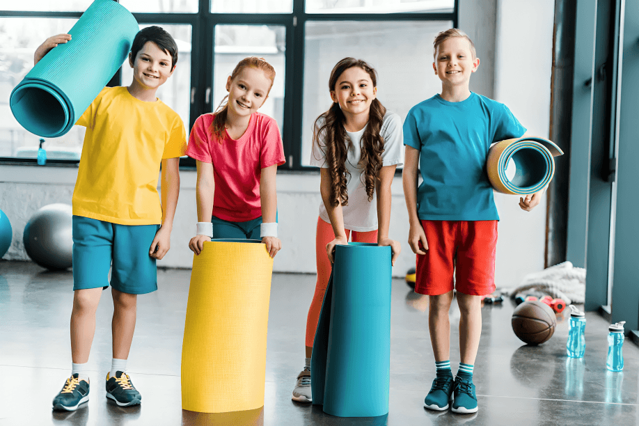 Four diverse children holding yoga mats in a gym.