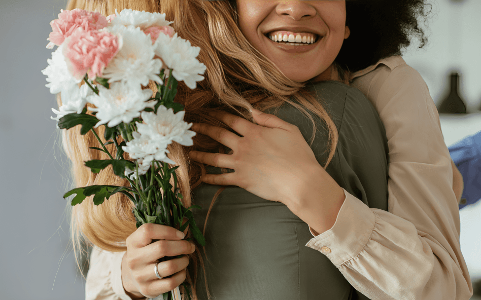 A smiling woman hugs another, holding pink and white flowers.