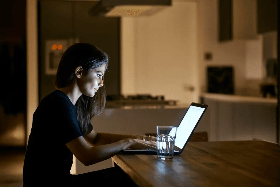 A woman works on a laptop at a wooden table at night.