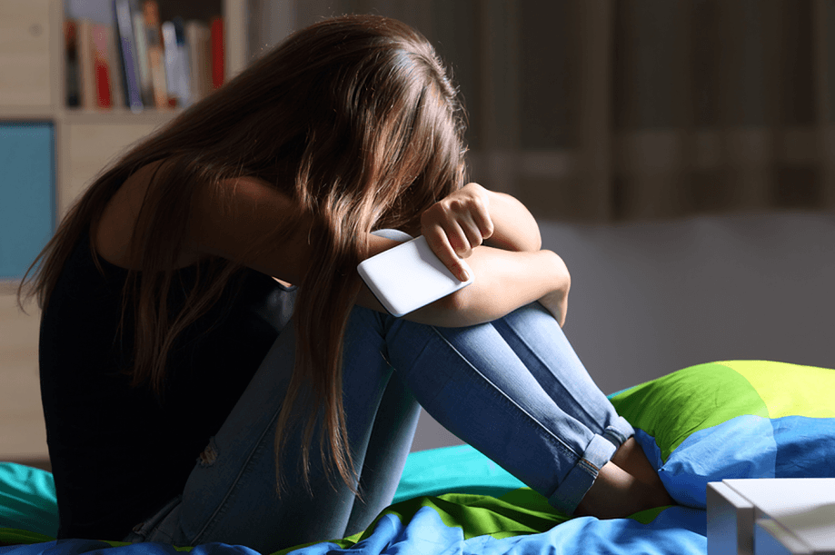 A young person seated on a bed holding a phone, possibly cyberbullying.