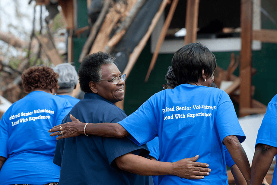 Senior volunteers survey disaster aftermath, supporting each other.