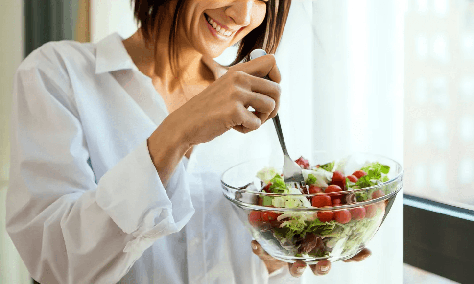 Smiling woman eating a fresh salad by a window.